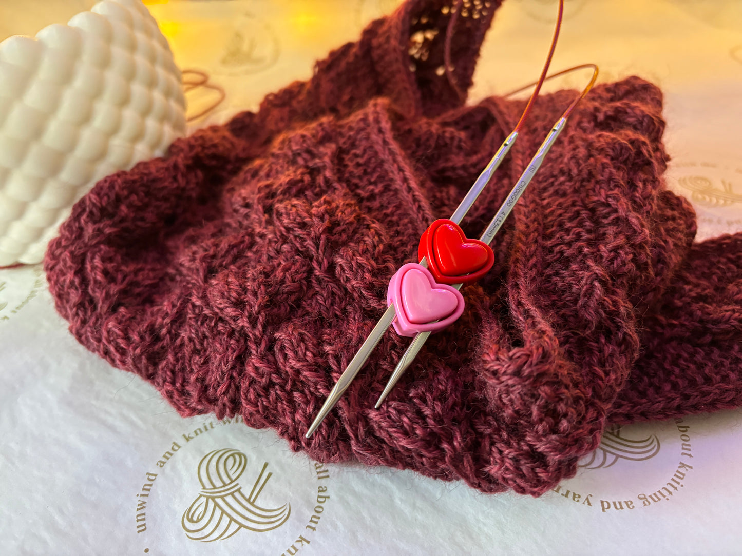 A close-up of a chunky knitted burgundy scarf or cowl with silver knitting needles resting on top, decorated with two heart-shaped pins in pink and red. The background is a white textured surface with a warm, soft-focus yellow light.