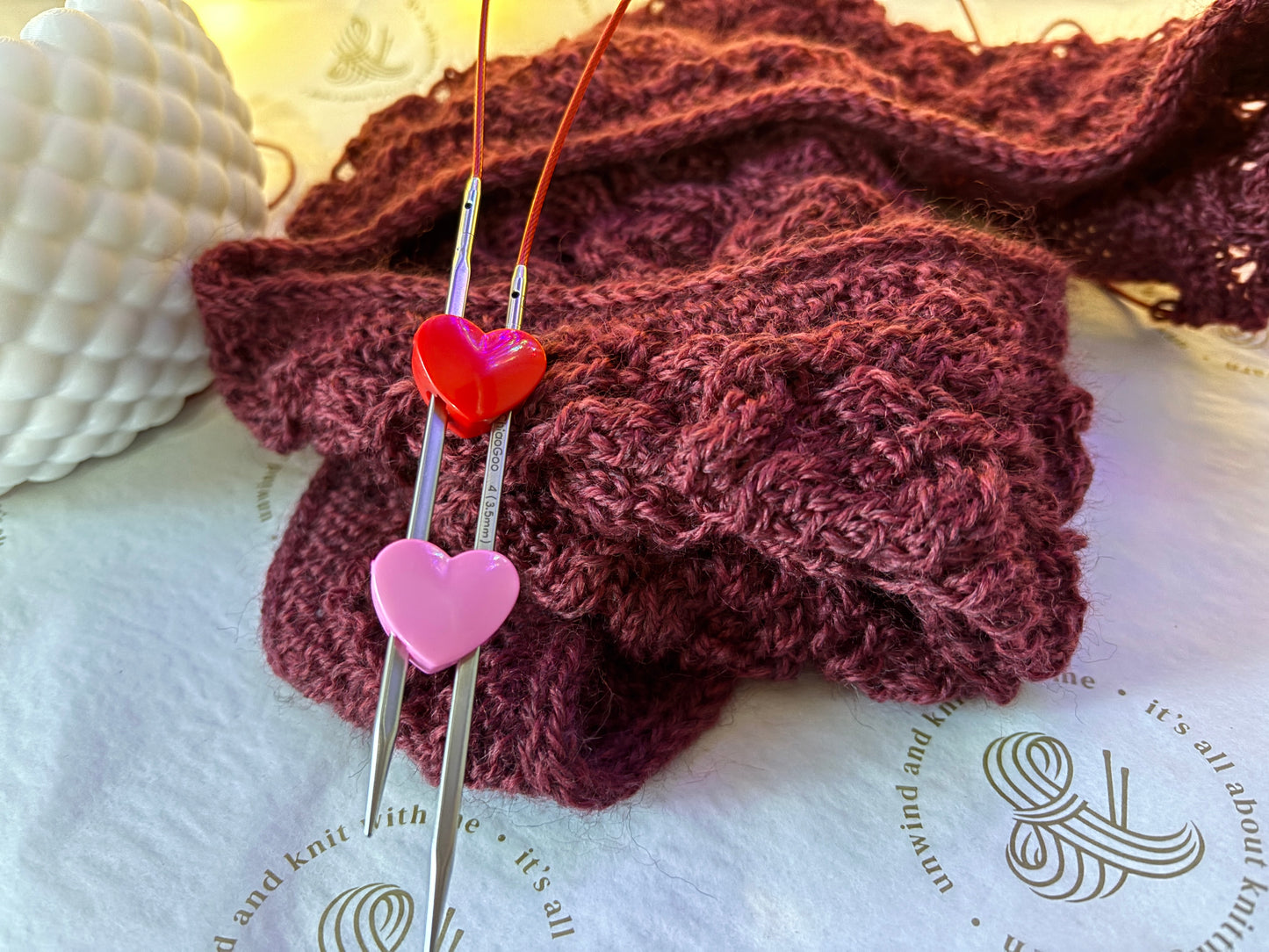 A close-up of a knitted burgundy scarf or cable-knit fabric draped on a white surface, with two metal knitting needles threaded through it and decorated with a red heart charm above a pink heart charm.